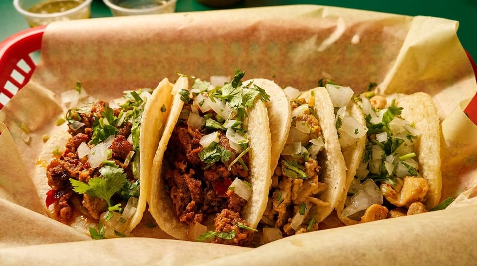 Four street tacos with various fillings, cilantro, and diced onions in a red basket.