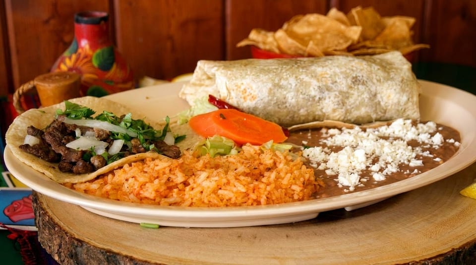 A plate with a burrito, a steak taco, rice, and refried beans topped with cheese.