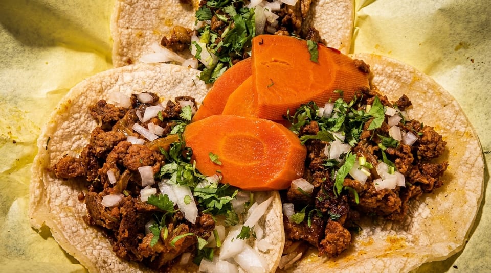 Three tacos with seasoned meat, diced onions, cilantro, and sliced carrots on yellow paper.