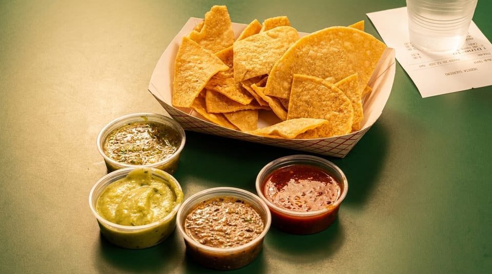 A basket of tortilla chips with four small containers of various salsas and guacamole.